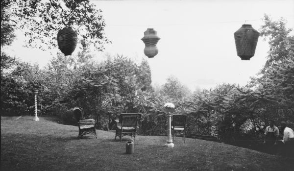 Japanese lanterns have been strung in preparation for the supper celebrating the wedding of Gertrude Merker and Clifford F. Messinger, which took place July 21, 1917.  Three wicker chairs and a glass gazing ball on a pedestal stand on the lawn which slopes away from the camera. There are two workmen at lower right.