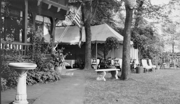 In preparation for the supper to celebrate the wedding of Gertrude Merker and Clifford F. Messinger, tables with tablecloths and chairs have been placed in a large tent near the porch of a house. An American flag is flying from a pole on the corner of the porch. There is a large house in the background. Chairs and a concrete bench are sitting on the lawn at right. In the foreground is a birdbath.    