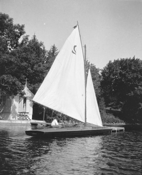 View across water towards an unidentified man wearing a cap sitting on the deck of a sailboat, the <i>Vidette</i> at the pier of Villa Henrietta, the Brumder family summer home at Pine Lake. The boathouse with elaborate gable fretwork is in the background. There is a woman standing in the open doorway of the boathouse.