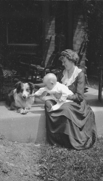 Margaret Bouer (Mrs. Herbert P.) Brumder holding her year-old son, Herbert Edmund Brumder, on her lap as he is reaching towards the family dog sitting on the left. They are on the porch of their brick home at 2030 E. Lafayette Place.