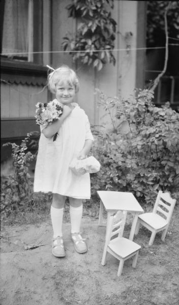 Barbara Brumder (1918-2017) posing on her fourth birthday with a bouquet of flowers tucked in her right arm. She is holding two packages in her left hand and is standing beside a doll-sized table with two chairs. She is wearing a short light-colored dress, long stockings and summer shoes.   