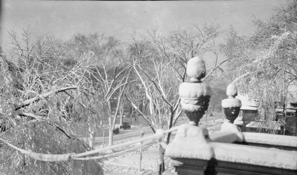 An elevated view of Grand (now Wisconsin) Avenue near 18th Street from the second floor of the home of Henriette (Mrs. George) Brumder. Snow is covering the ground and clinging to tree branches and utility wires. Two snow-covered stone urns decorate the roof over the front porch. There are two automobiles parked on the street.