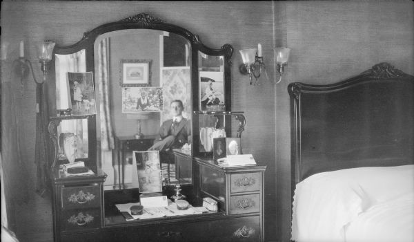 Herbert Brumder is reflected in the center mirror of a vanity in the bedroom of his home at 2030 East Lafayette Place. Partially obscuring the mirrors are snapshots of his wife, Margaret, and their three children, Herbert Edmund, Barbara, and Philip. Wall sconces fitted for both gas and electricity flank the vanity, and neckties are hanging from the sconce on the left. The bed is on the right.