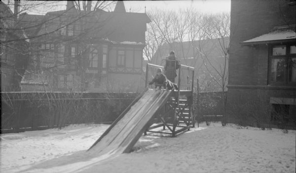 Barbara and Herbert Edmund, the two older children of Herbert P. and Margaret Bouer Brumder, posing with their sleds at the top of a wooden slide in the backyard of their home at 2030 East Lafayette Place. They are bundled in winter coats, hats and scarves, and snow is on the ground. The large house next door has half-timber detailing on the second floor.