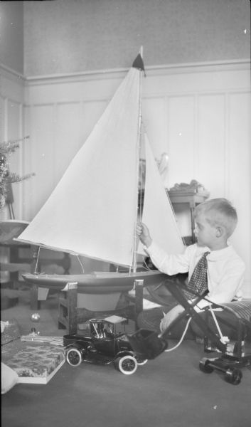 Herbert Edmund Brumder is sitting on the floor holding the halyard of a large toy sailboat which is resting on a stand. On the floor in front of him are a toy pickup truck and dragline. A few branches of the Christmas tree are on the far left.
