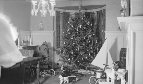 A large Christmas tree fills the bay window in the dining room of Herbert P. and Margaret Brumder. There are Christmas gifts arranged around the tree, and include a child's winter coat and hat, two child's quadricycles, a toy dog-drawn milk cart, large sailboat, and a pickup truck. A note below the sailboat reads, in part: "This I give Herbert for good arithmatic[sic] and ..." At left, a man's torso partially obscures the flashlight source. 