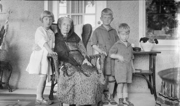 Henriette Brandhorst Brumder (1841-1924), the widow of Milwaukee publisher and businessman George Brumder (1839-1910) poses while sitting in a wicker chair, with grandchildren, from left, Barbara, Herbert Edmund, and Philip George Brumder. They are the children of Henriette's youngest child, Herbert Paul Brumder and his wife Margaret (Bouer). They are on the porch at the Brumder family summer home, Villa Henrietta, at Pine Lake.   