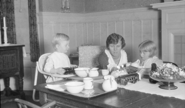 Philip George Brumder, sitting in a high chair on the left, and his sister Barbara, sitting on the right, watch intently as their mother, Margaret Bouer (Mrs. Herbert P.) Brumder tends the three lighted candles on Philip's small birthday cake. There are enamelware dishes on the table and a centerpiece of cut flowers. Philip is holding a toy locomotive on the tray of his high chair; other train cars are on the table. They are in the dining room of the family's home at 2030 East Lafayette Place.