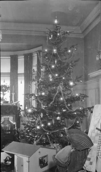 A tall, lighted Christmas tree stands in the dining room of the Herbert P. Brumder family residence, 2030 East Lafayette Place. Glass ornaments and tinsel decorate the tree, and there are toys arranged in front of it. These include a garage for a doll house, a toy baby buggy and a quilt.
