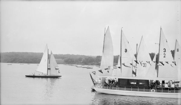 Elevated view of a motor launch, "dressed" with signal flags, alongside several racing yachts on a lake.  