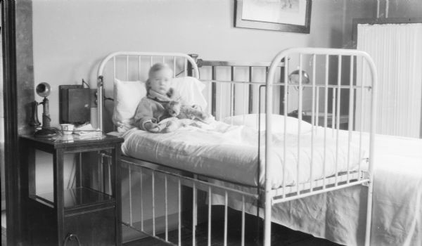 Philip George Brumder, son of Herbert Paul and Margaret Bouer Brumder, resting in a hospital crib holding a stuffed dog and a doll. His cheeks are swollen. There is a telephone and an enamelware cup on the bedside table. An adult sized bed is next to the crib, and there is a fabric covered portable screen in the background on the right.