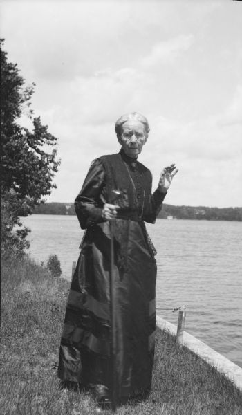 Henriette Brandhorst Brumder (1841-1924), widow of publisher and businessman George Brumder (1839-1910), standing on the shore of Pine Lake. She is wearing a dark, long dress and is holding a walking stick. There is a stone or concrete seawall along the shoreline on the right.