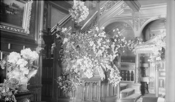Multiple bouquets and other flower arrangements fill the entry hall of the George and Henriette Brumder mansion at the corner of Grand (later Wisconsin) Avenue and 18th Street, on the occasion of Henriette's death, April 19, 1924. Elaborately carved wood paneling covers the walls and and extends up the grand stairway.  