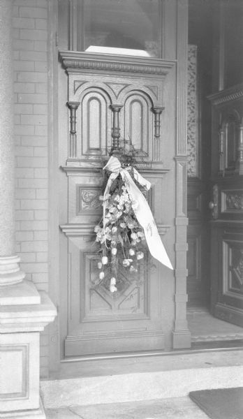 A floral spray with large ribbon hangs on the left door of the front entry of the George and Henriette Brumder mansion at the corner of Grand (later Wisconsin) Avenue and 18th Street, on the occasion of Henriette's death, April 19, 1924. The doors are richly carved. There is a stone column at far left.
