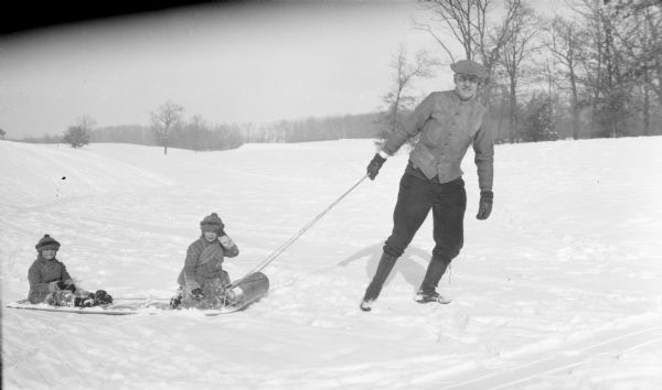 Herbert Paul Brumder is pulling his two sons, Herbert Edmund, center, and Philip George, left, on a toboggan in a snowy, rolling landscape. The two boys are wearing knit hats and heavy coats.