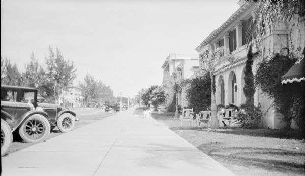 Automobiles are parked along Lincoln Road in front of the Spanish Colonial Revival style Lincoln Hotel. Two men are sitting in chairs which have the hotel's name painted on the back. Small palms and other trees line the street.