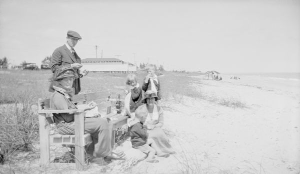 Margaret Bouer Brumder is sitting on a log on a beach, with her son Philip beside her. Her other children, Barbara and Herbert Edmund are standing behind them. An unidentified woman is sitting on a bench at left; a man on the left is standing behind the bench. There are two bathhouses in the background, and open shelters are on the beach near the water. A line of utility poles stretches into the distance.  