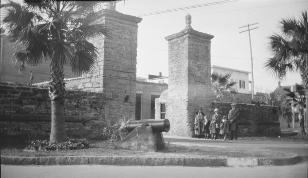 Margaret (Mrs. Herbert P.) Brumder, center, posing with her children, from left, Herbert Edmund, Philip, and Barbara. Barbara is holding the hand of an unidentified man standing on the right. There is an unidentified woman on the far left. The group is standing in front of one of the two stone towers of St. Augustine's old city gate. There is a cannon in the foreground.