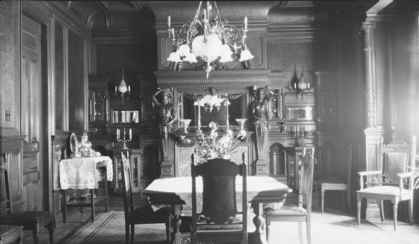 A view of the dining room in the George and Henriette Brumder mansion at the corner of Grand (later Wisconsin) Avenue and 18th Street. Sunlight bathes the room from windows beyond the right frame of the image. The fireplace is flanked by an Atlas, left, and a caryatid. There is a silver coffee and tea service on a small table against the wall on the left. A vase of flowers is standing on the dining table. The chandelier and sconces are fitted for both gas and electricity.