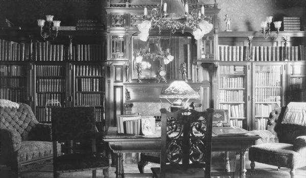 Tall bookcases flanking a large fireplace in the library of the George and Henriette Brumder mansion at the corner of Grand (later Wisconsin) Avenue and 18th Street. The overhead chandelier has fittings for both gas and electricity. A Tiffany style lamp with poppy shade is standing on the sturdy library table. There are desk chairs at the table and an upholstered easy chair on either side.