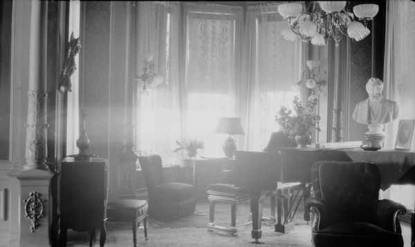 A sculpted bust of George Brumder (1839-1910) is sitting on a grand piano near a bay window in the Brumder mansion at the corner of Grand (later Wisconsin) Avenue and 18th Street. The chandelier and wall sconces have been fitted for both gas and electricity. There is a wooden music stand at left. On the piano is a flower arrangement, and another  one is on a small table near the window.