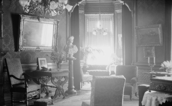 A parlor or sitting room, with a view into a small room beyond a doorway, probably in the tower of the George and Henriette Brumder mansion at the corner of Grand (later Wisconsin) Avenue and 18th Street.  There is damask style wall covering above textured and patterned wainscoting. The chandelier in the far room has been fitted for both gas and electricity. A sculpted bust of a muse with a lyre stands at the left of the doorway. There are upholstered chairs in both rooms.