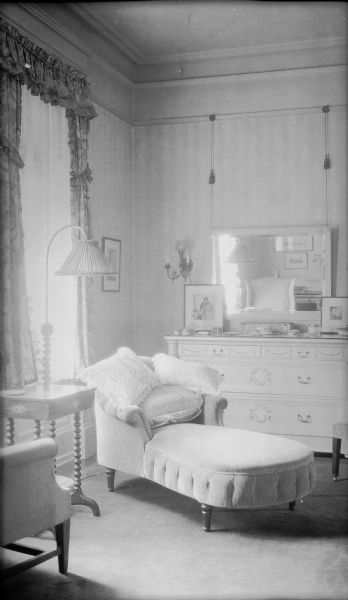A floor lamp is standing on the left beside a chaise lounge in a bedroom of the George and Henriette Brumder mansion at the corner of Grand (later Wisconsin) Avenue and 18th Street. There is a dresser against the far wall; a mirror is hanging above it on long cords attached to a picture rail. There is a small table and wing back chair on the left.