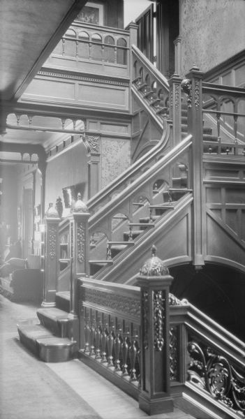 A view of the carved wooden staircase taken from the second floor landing in the George and Henriette Brumder mansion at the corner of Grand (later Wisconsin) Avenue and 18th Street. There is wood paneling and patterned wallpaper in the stairwell. In the background is an upholstered settee.