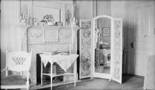 A small table with books on the lower shelf is standing in front of the fireplace in a bedroom of the George and Henriette Brumder mansion at the corner of Grand (later Wisconsin) Avenue and 18th Street. There is a folding screen to the right of the fireplace with a central mirror and fabric side panels.