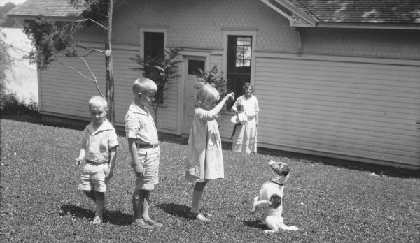 A small, wet dog "sits up" at the command of Barbara Brumder as her brother Herbert E. (center) are looking on. Younger brother Philip George, left, is looking at the camera. The children's mother, Margaret (Mrs. Herbert P.) Brumder is looking on from the background, while standing next to the boathouse. The boathouse was part of the original George Brumder summer estate, Villa Henrietta, on Pine Lake.