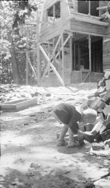 Philip George Brumder, the youngest child of Herbert Paul and Margaret Brouner Brumder, is picking up a brick from a pile on a construction site. The house under construction in the background has wood sheathing over the frame, and boards supporting the structure.