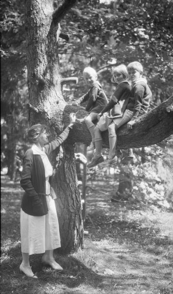 Margaret Brouner (Mrs. Herbert P.) Brumder, left, standing near the trunk of a large tree. Her three children, from left, George Philip, Barbara and Herbert E., are sitting on a large limb of the tree. They are wearing sweaters over casual clothes.