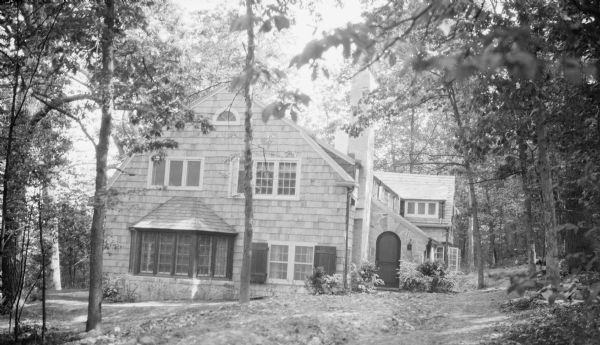 Exterior view of the new shingle style lake house belonging to Herbert P. and Margaret Brumder. A tall stone chimney rises near the main entrance, which features a door with an arched top, set into a stone wall. There is a large bow window on the side. The lot is heavily wooded. Pine Lake is down the hill to the left.