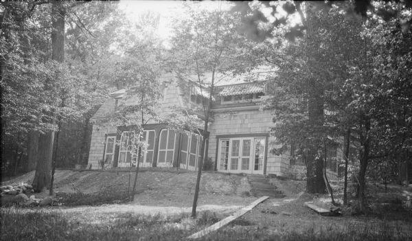 A view of the east, lake-facing facade of the new shingle style house of Herbert P. and Margaret Brouner Brumder. There are awnings over the second story windows. French doors at right lead to steps down the sloped lawn. A gabled center wing features a screened porch on the ground level. The lot is heavily wooded.