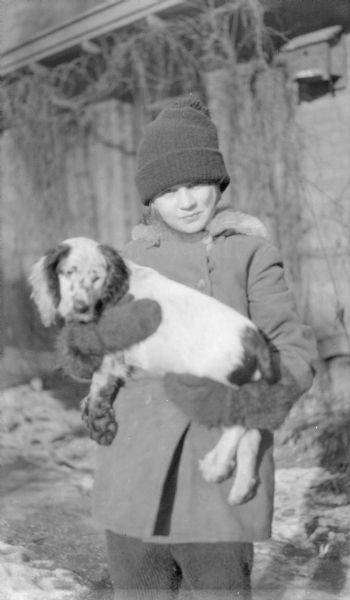 Barbara Brumder, the daughter of Herbert Paul and Margaret Bouer Brumder, holding a puppy, probably a Setter or Spaniel. She is wearing a stocking cap, coat and mittens. In the background is a birdhouse mounted on a building, and leafless vines covering a trellis supported by the overhang of the building.  