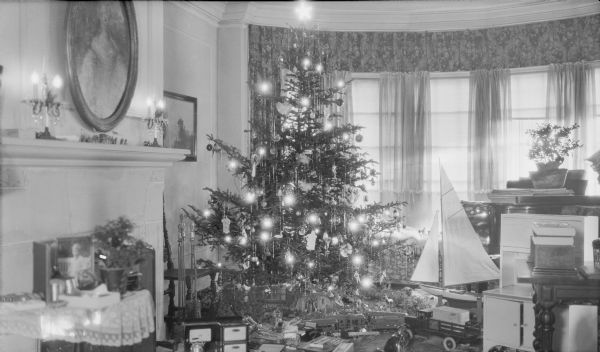 An electrically lit Christmas tree standing near the bow window in the living room of Herbert and Margaret Brumder, 2030 East Lafayette Place. An abundance of toys surround the tree, including a toy kitchen cabinet and cook stove, sailboat, truck, and train set. At left is a fireplace with two electric candelabras on the mantle.