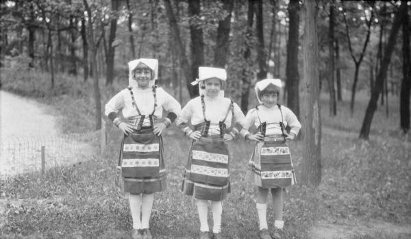 Barbara Brumder, left, posing with her cousins Mary Baldwin Messinger, center, and Joanne Beecher Messinger, right. The girls are wearing similar ethnic German costumes, or <i>Tracht</i> which include white headdresses, suspenders, and embroidered aprons. They are in a wooded area, and there is a low wire fence on the left.