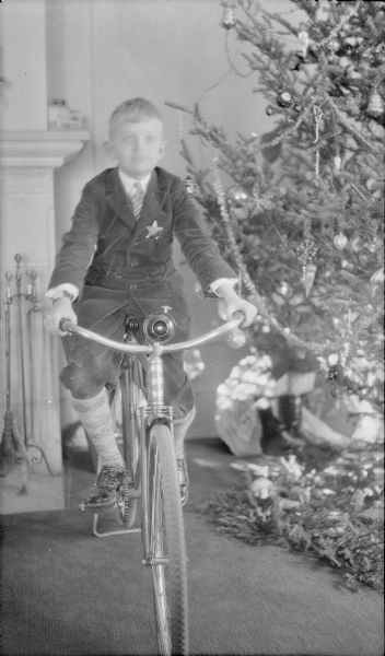 Herbert Edmund Brumder posing on his bicycle next to the Christmas tree in the family living room at 2030 East Lafayette Place. He is wearing a suit with knickers and has a star shaped badge on his jacket. The corner of the fireplace is in the background on the left.