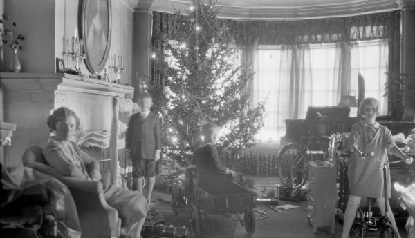 Margaret Bouer (Mrs. Herbert Paul) Brumder, left, is sitting in an armchair in the living room of the family home at 2030 East Lafayette Place. Her children, from left, are Herbert Edmund, Philip George, and Barbara, who are posing surrounded by their Christmas gifts. Philip is sitting in a pedal truck and Barbara is on her bicycle. There is a larger bicycle in the background, near a grand piano. A large Christmas tree with electric lights is standing near the bow window. A cutout Santa Claus is peering from behind the corner of the fireplace.