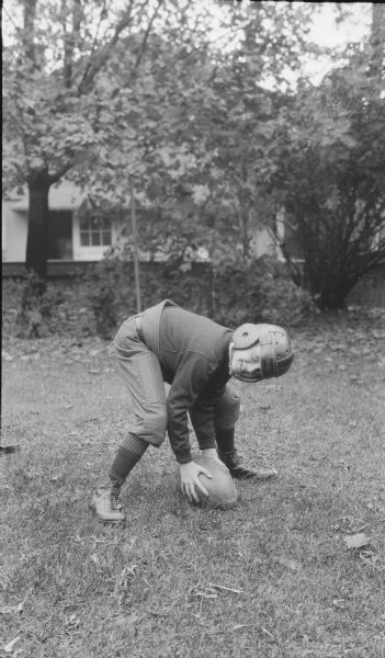 Herbert Edmund Brumder posing outdoors as a center ready to snap his football. He is wearing a leather helmet, football jersey and padded football pants. There is a building in the background.
