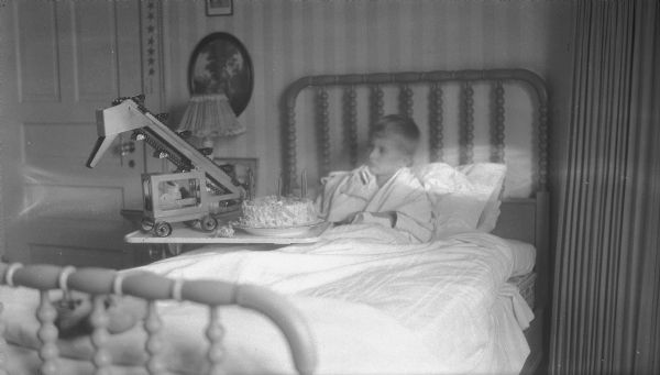 Philip George Brumder is sitting propped up in a single bed. He is looking at a toy sand or gravel conveyor which is standing next to a birthday cake with six candles on an overbed table. There is also a small toy pig on the table.