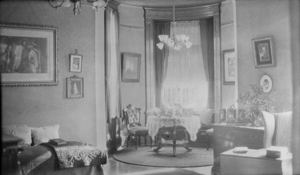 An interior view of the George and Henriette Brumder residence at the corner of Grand (later Wisconsin) Avenue and 18th Street. The view is from a large room into a small, circular room with tall windows.  There is a window seat under the far window. A chandelier in the small room is fitted for both gas and electricity. Below it, a potted vine is sitting on a round table. Framed prints and photographs are hanging on the walls.
