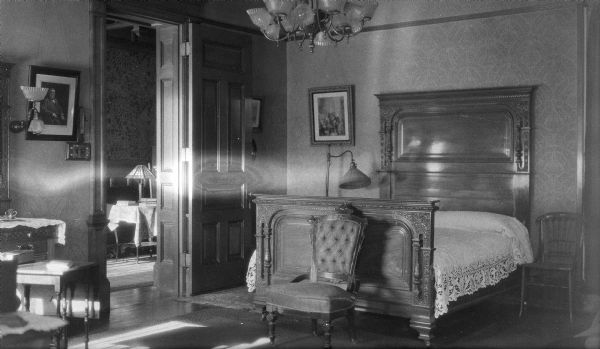 A view of a bedroom in the George and Henriette Brumder mansion at the corner of Grand (later Wisconsin) Avenue and 18th Street. The bed has a tall carved headboard and shorter foot; a lace bedspread covers the mattress and pillows. A wall sconce at left, fitted for both gas and electricity, matches a large overhead chandelier. A framed photograph of George Brumder is hanging near the sconce. There is a table with books and a table lamp visible through the doorway.
