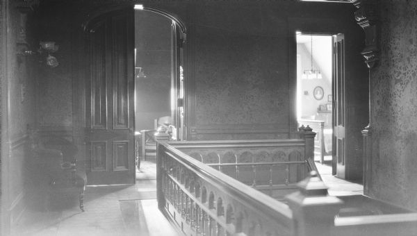 A view of the third floor landing of the main stairway in the George and Henriette Brumder mansion at the corner of Grand (later Wisconsin) Avenue and 18th Street. There is a carved wooden corbel at upper right. At left, one of the doors of an arched double doorway provides a glimpse into the room beyond. A smaller doorway at right leads into a bedroom.