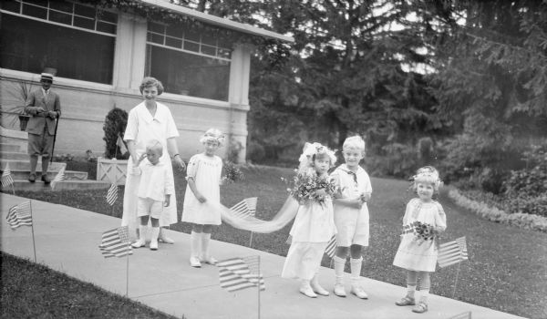Clifford Messinger, rear left, and his wife, Gertrude Merker Messinger supervising a mock wedding at Villa Henrietta at Pine Lake. The bride is their daughter, Mary Baldwin Messinger; another daughter, Joanne Beecher Messinger is the flower girl, far right. The groom is Herbert Edmund Brumder; his sister Barbara is holding the bride's train. Philip George Brumder is standing near Gertrude. The wedding party is standing on a sidewalk lined by small American flags. Mary, the bride, is carrying a large floral bouquet, and all three girls have flowers in their hair.