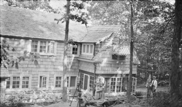 Four children posing in the yard of a new, two-story shingle style house belonging to Herbert Paul and Margaret Bouer Brumder. Near a hand pump, center, Barbara Brumder is embracing her first cousin once removed, Mary Messinger. Philip and Herbert E. Brumder are standing at far right; Philip is holding an American flag.