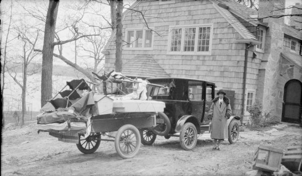Margaret Bouer (Mrs. Herbert Paul) Brumder posing standing beside an automobile hitched to a trailer, which is precariously loaded with household furnishings including shelves, chairs, mattresses, and a dog house. The car is parked near the front door of the Brumder's new two-story shingle style house. Margaret is wearing a hat, and a coat with a fur collar. Pine Lake can be seen through the leafless trees at left.  