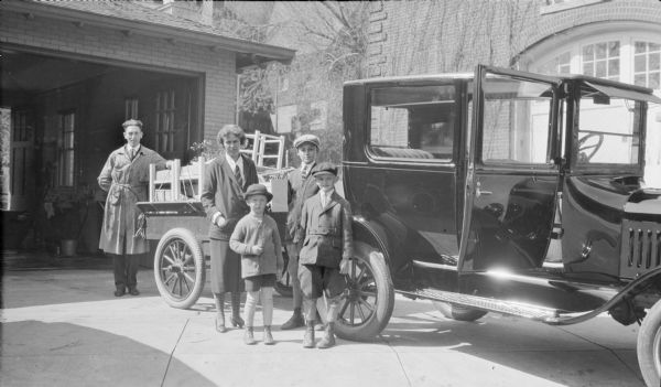 Margaret Bouer (Mrs. Herbert P.) Brumder (center) is standing with her sons Philip (front, left) and Herbert E. (front, right) near a trailer hitched to an automobile. The trailer is loaded with furniture. The man standing at left and the boy behind Herbert are not identified. There is a brick garage in the background, and the garage of another house is at right.