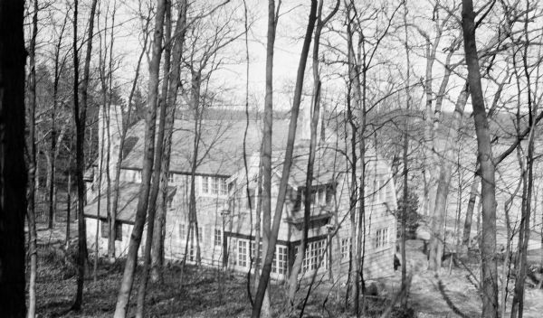 An elevated view looking down the hill through the leafless branches of trees towards the newly constructed shingle-style house belonging to Herbert Paul and Margaret Bouer Brumder. The two-story house has two large stone chimneys, the one at left near the shed roofed entryway.  The lake is in the background on the right. A wing extending from the rear of the house has a screened sleeping porch above a sunroom.  