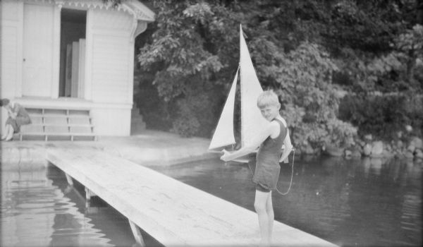 Herbert Edmund Brumder, wearing a bathing suit, is standing on a wooden pier holding a large toy sailboat. An unidentified woman is sitting on the steps of the bathhouse in the background. They are at Villa Henrietta, the Brumder family estate on Pine Lake.